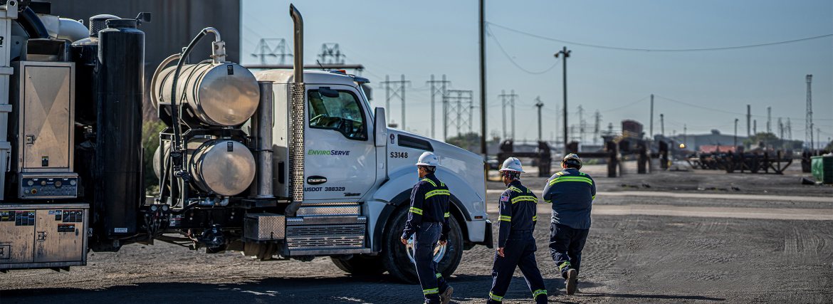 pictured: three EnviroServe workers wearing coveralls and hardhats walk near a large white truck