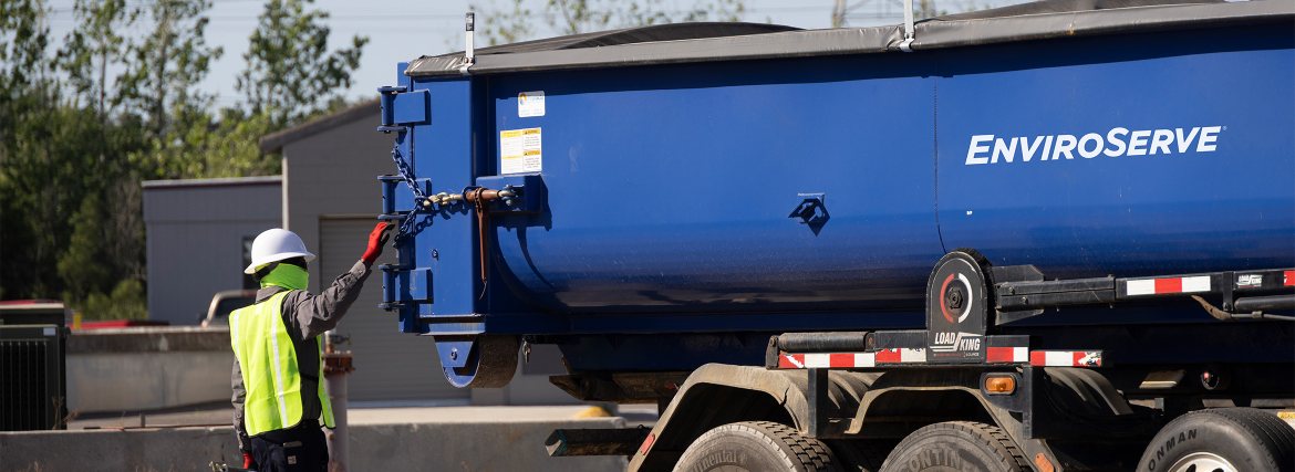 pictured: an EnviroServe waste management worker wearing a safety vest and hardhat stands next to a blue truck bed