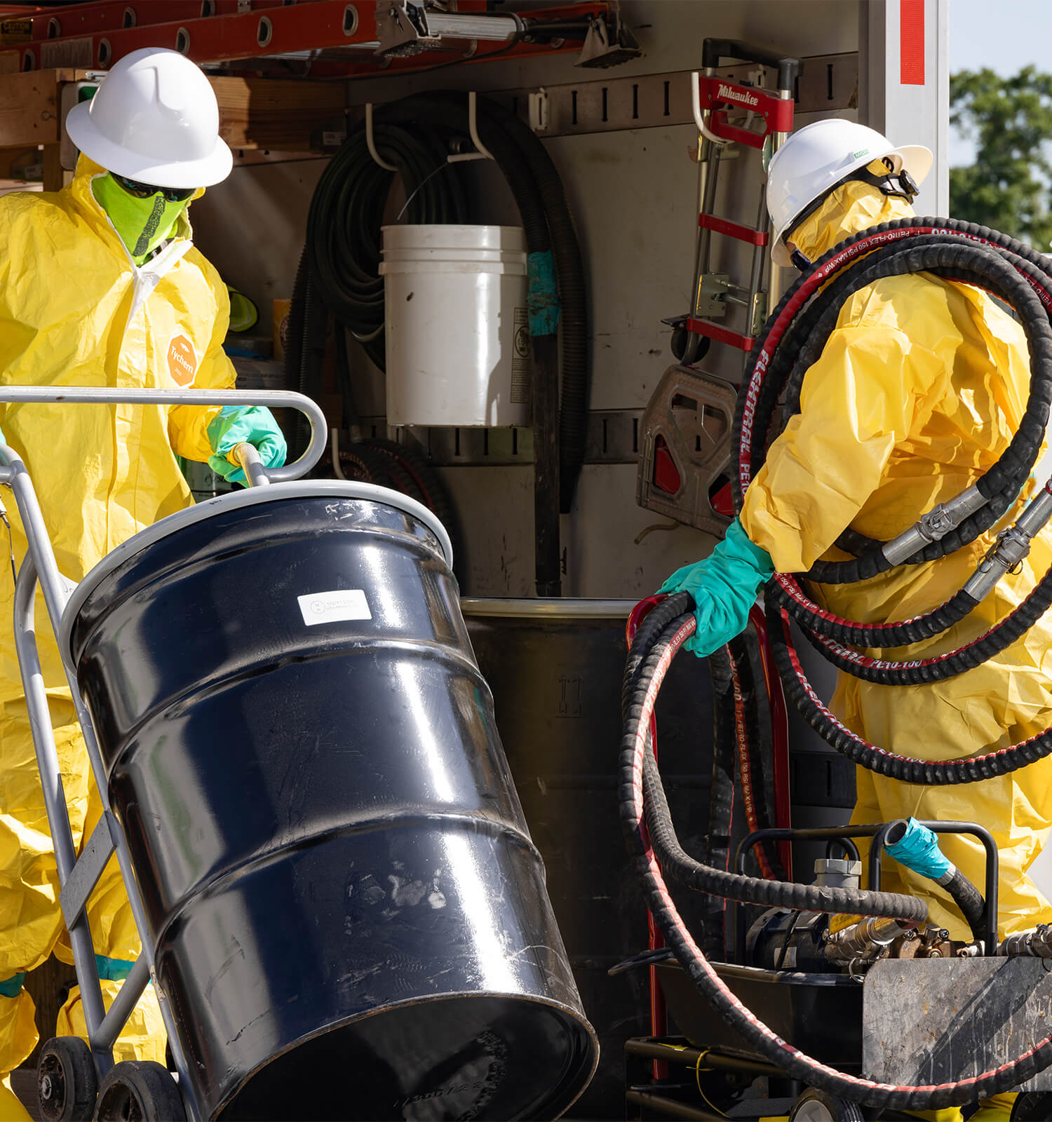 pictured: two EnviroServe workers wearing masks, hard hats, and yellow hazmat suits move hoses and a barrel of hazardous waste