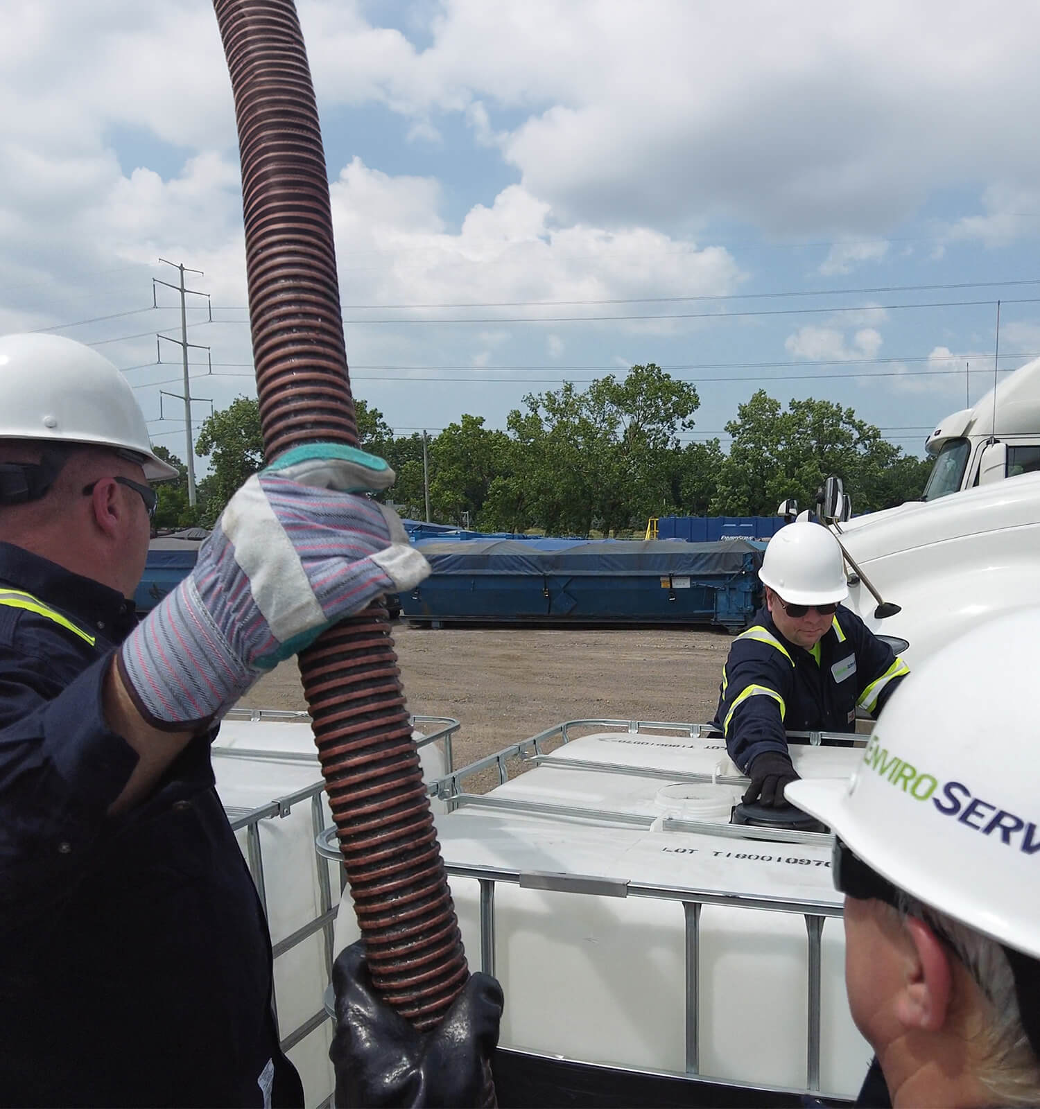 pictured: three EnviroServe workers wearing gloves, coveralls, and hardhats work with hazardous liquid