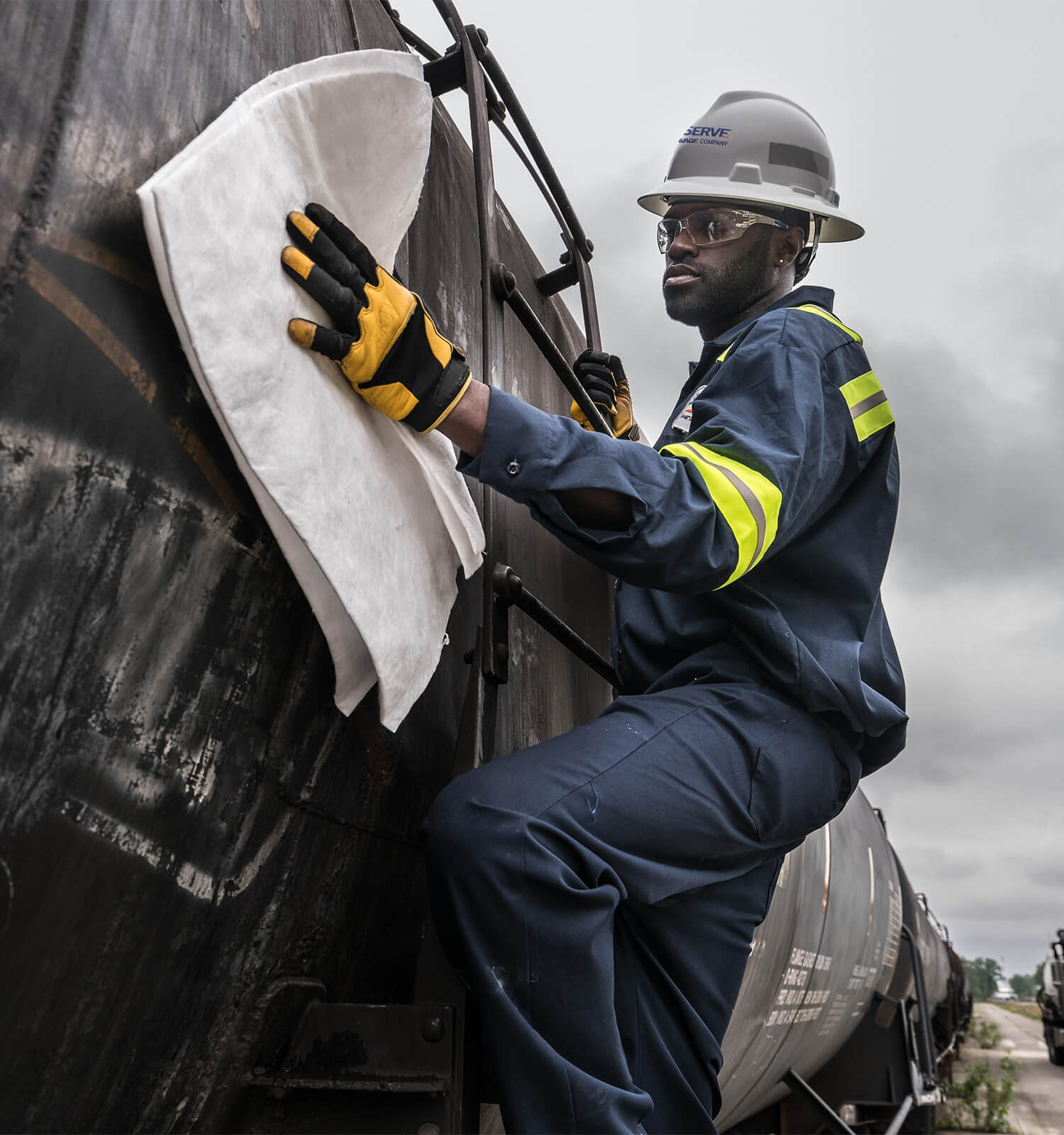 pictured: an EnviroServe worker wearing coveralls, safety goggles, safety gloves, and a hard hat cleans the side of a rail car