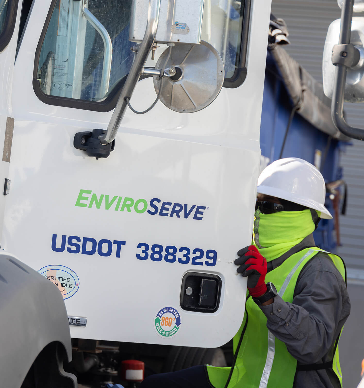 pictured: an EnviroServe worker wearing safety vest, safety gloves, mask, eye goggles, and hard hat opens the door of an EnviroServe semi truck