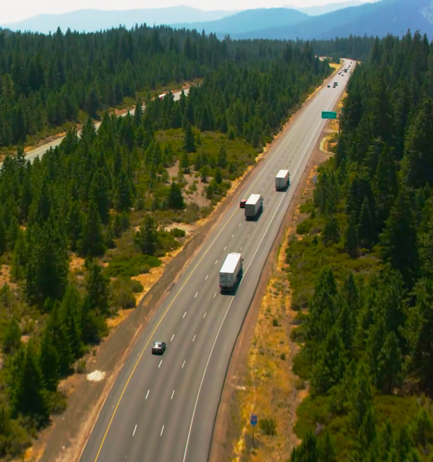 pictured: aerial photo of three EnviroServe waste handling trucks driving over a stretch of highway through coniferous mountain forest